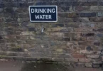 An outdoor drinking water tap with a sign on a stone wall in England