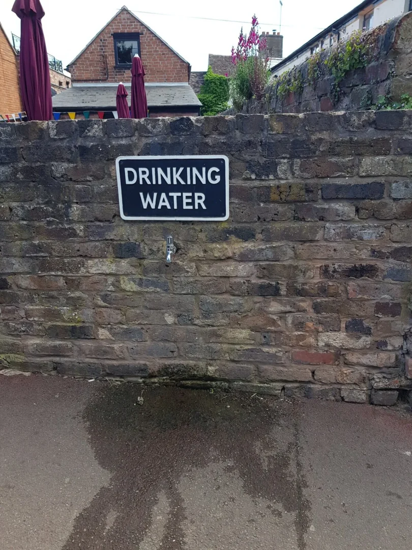 An outdoor drinking water tap with a sign on a stone wall in England